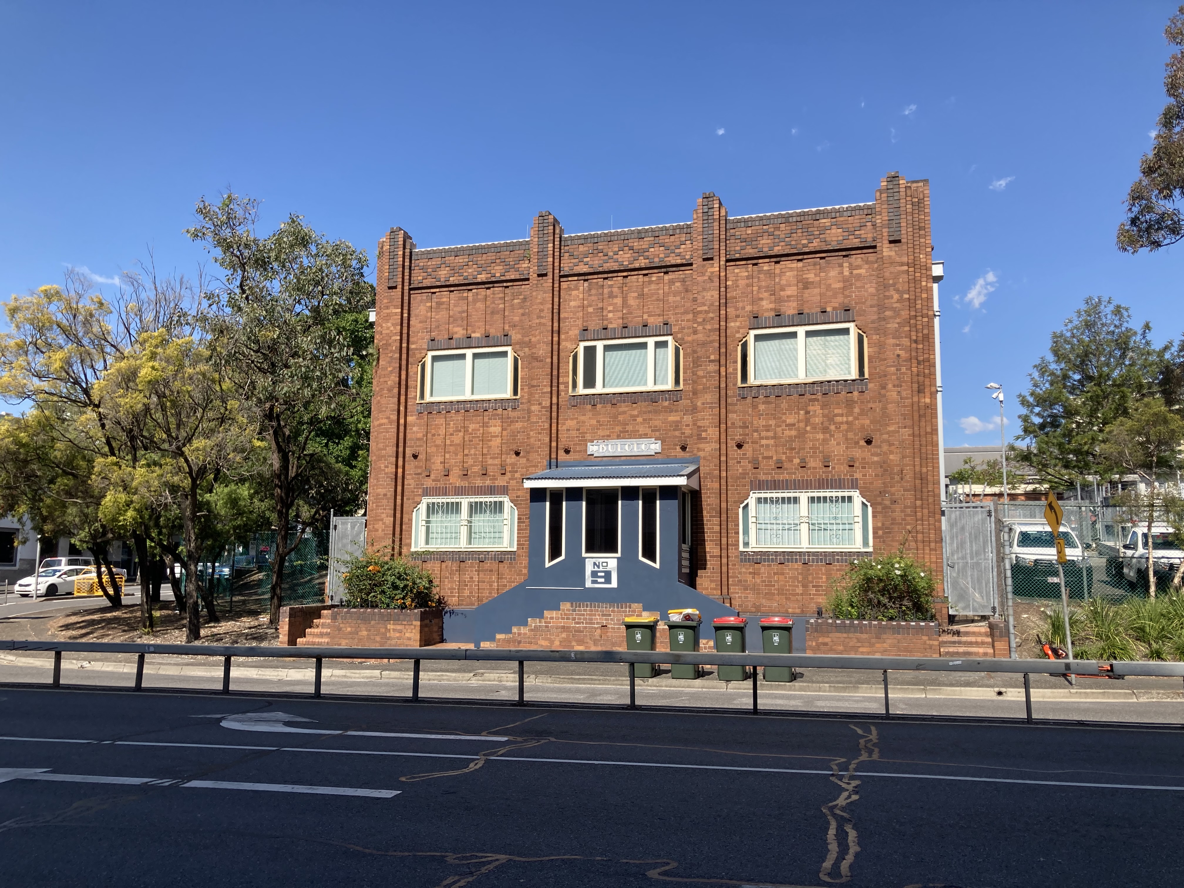 Two storey red brick building. A formal entrance is painted in dark grey. A wide bitumen road with a concrete central fence is in the foreground