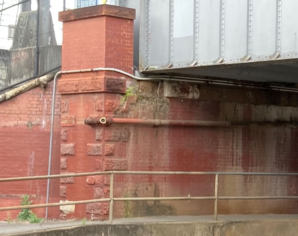 A red brick and stone abutment, supporting a steel bridge. A small amount of green vegetation is growing round the structure, and a concrete path with a metal fence is in front of the abutment.