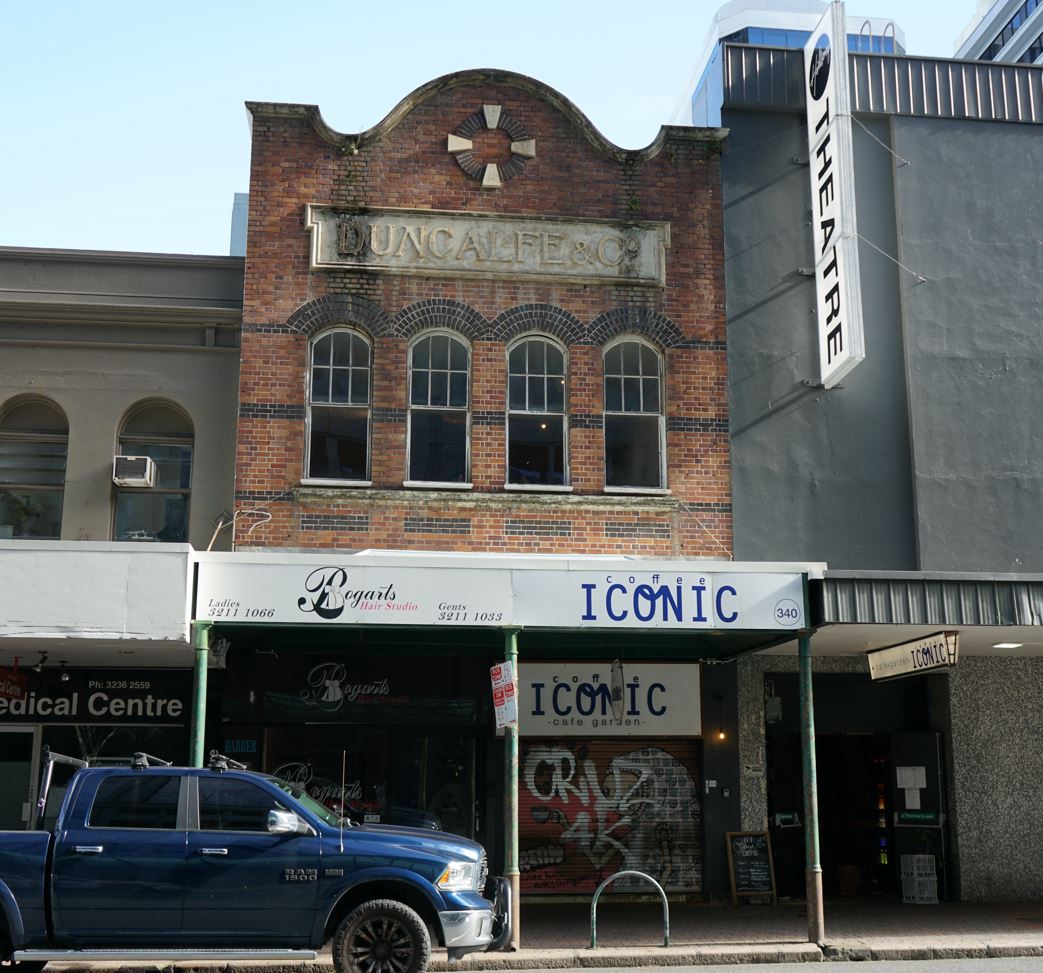 A two storey, brick building, with four windows on the top floor. Above the windows is a plaque reading "Duncalfe & Co". A modern awning is at the first floor, with "Bogarts" on the left and "Coffee Iconic" on the right. A bitumen road and truck are in the foreground. 