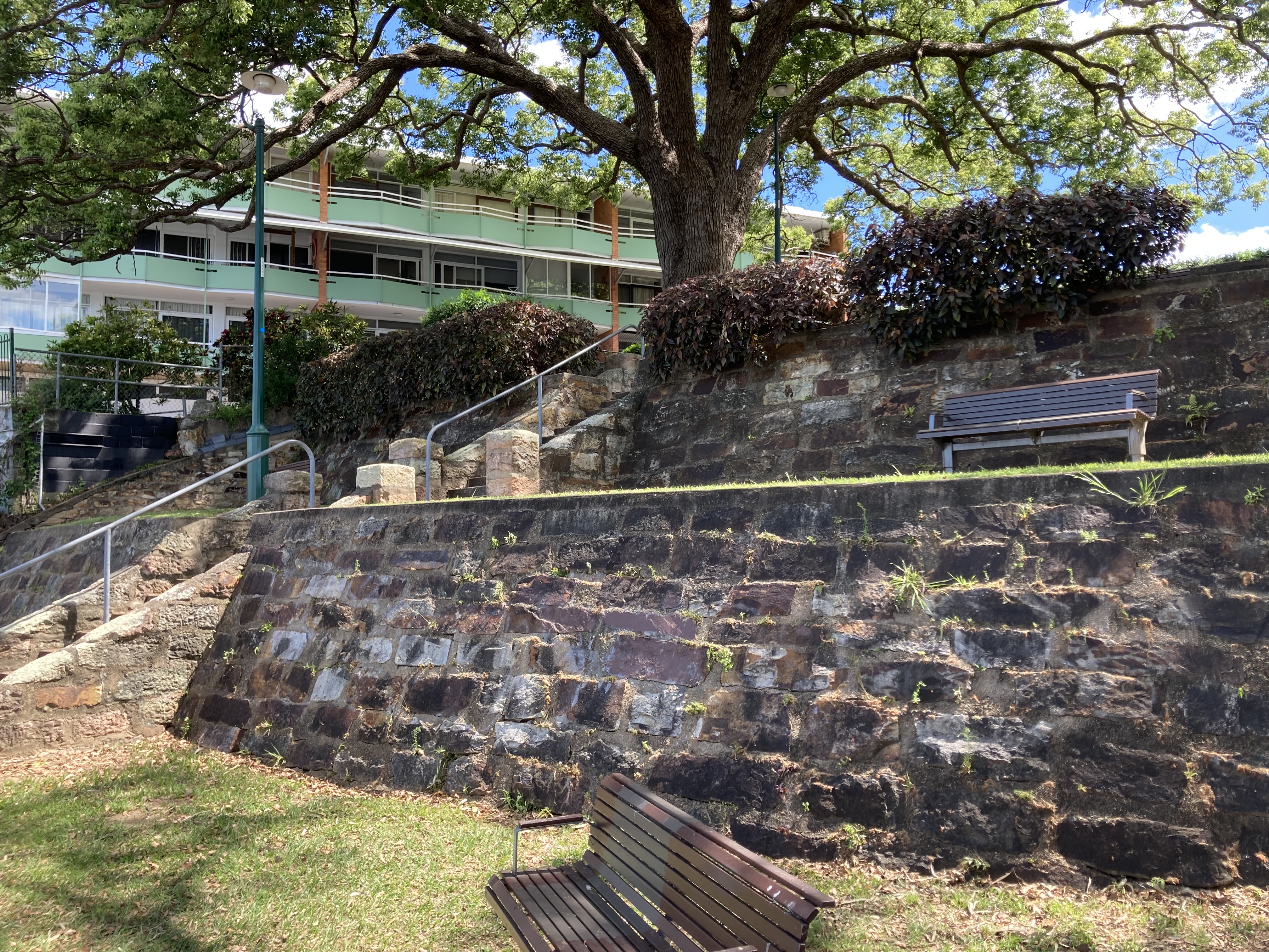 A large stone block wall. Stone stairs with a metal railing are in the background, with a large apartment block and mature trees. Small timber and metal benches are in the foreground and middle ground.