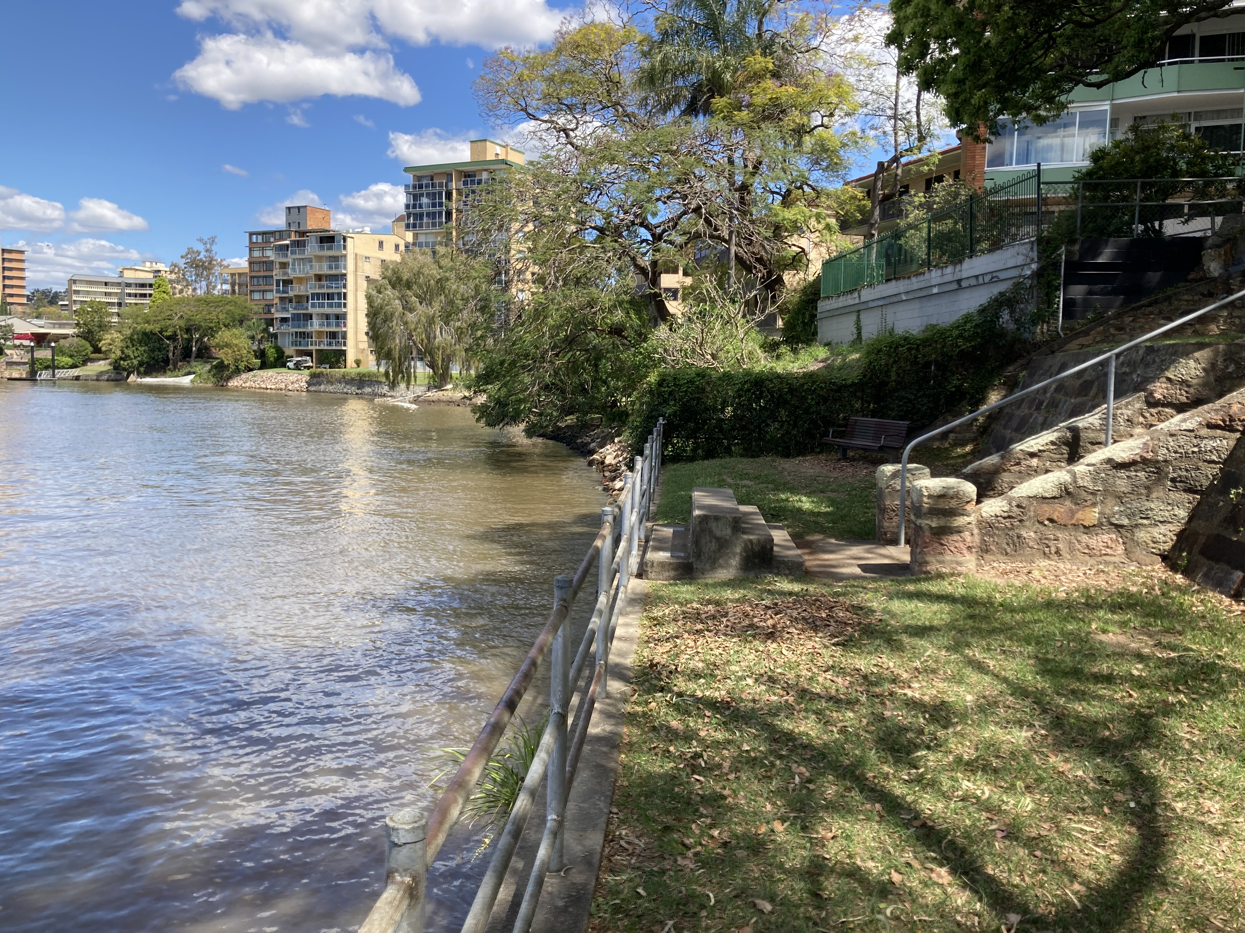 A park at the edge of the Brisbane River. The River is on the left of the image. A metal railing separates the grassy area of the park and the River. A small set of concrete stairs in the centre of the image lead up to the railing. A larger set of stone stairs continue off the right of the image. The background contains large apartment blocks and mature trees.