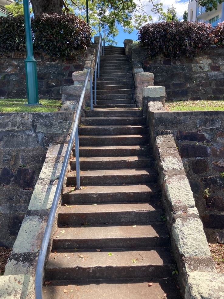 A set of stone stairs, with a metal railing. Short stone walls are located next to the set of stairs, with grass and other vegetation throughout the image.