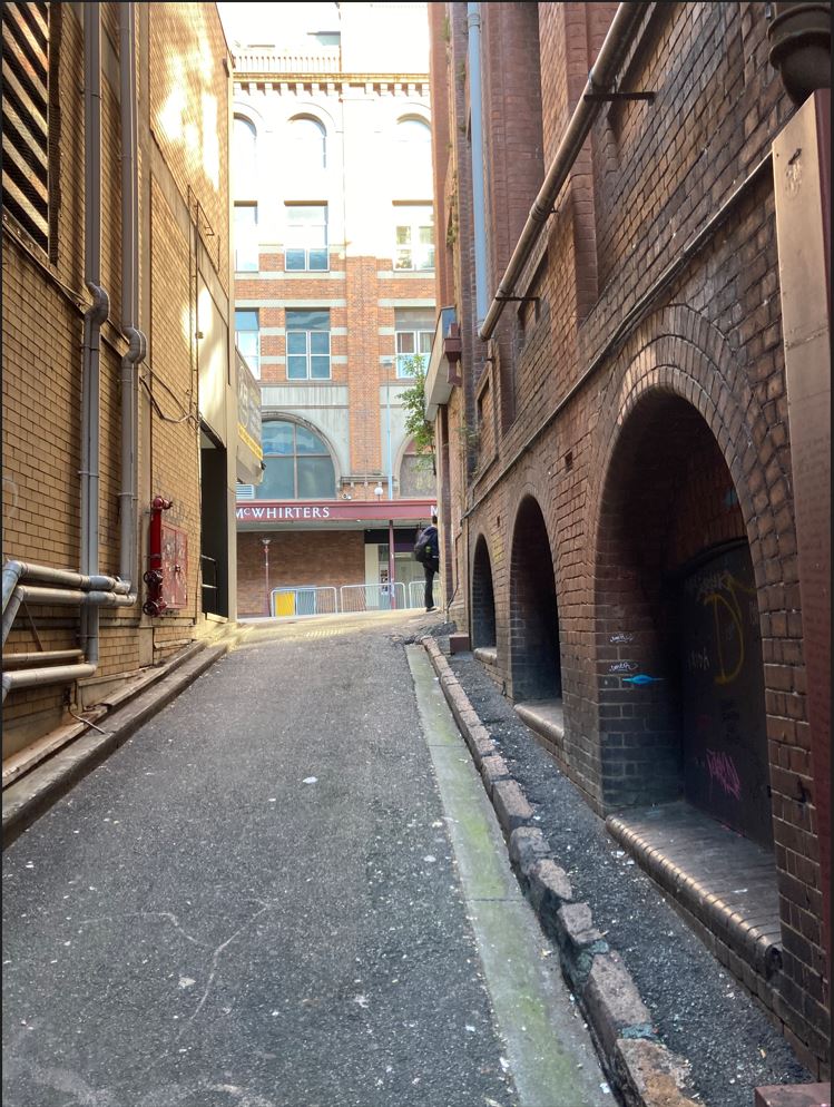 Overells Lane, a bitumen laneway between two brick buildings. Stone blocks are used for kerbing