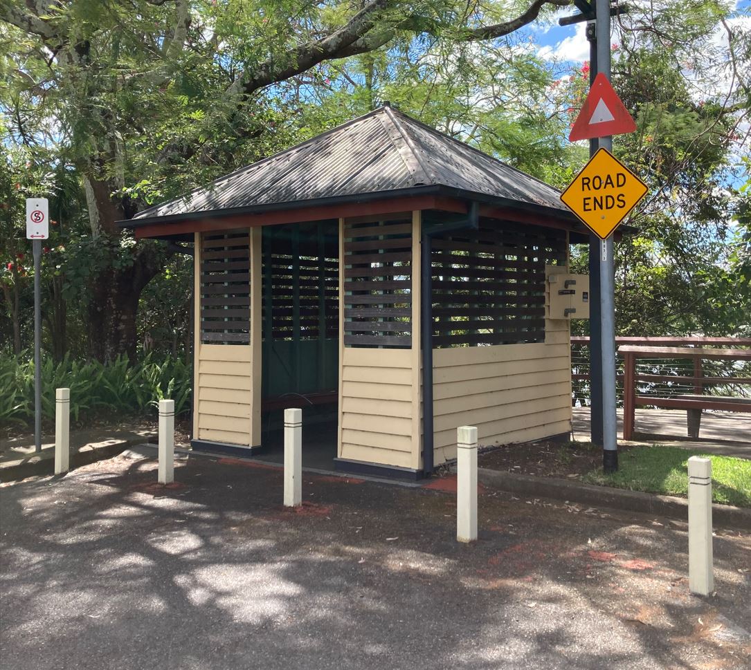 A single storey, timber ferry terminal, painted cream with blue trims. Mature trees are on either side of the structure. Warning, Road Ends, and No Standing signs are to the sides of the terminal building. A timber deck can be seen to the rear, and timber bollards are located at the front of the structure.