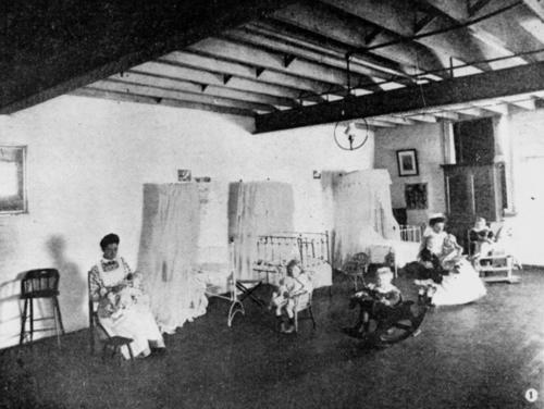 This is an image of ‘Interior view of the Brisbane Institute of Social Service, 1907', showing a couple of children are playing on rocking horses, while younger children are being nursed by kindergarten staff. Mosquito nets hang over three cots along the back wall.
