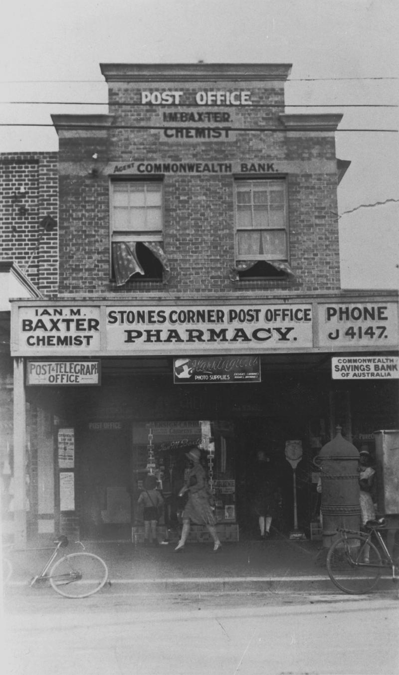 Two story brick Stones Corner Post Office, chemist and Commonwealth Bank building. People walk in front of hte building, and two upstairs windows are open. Advertisements for the business within are placed on the ground floor awning.