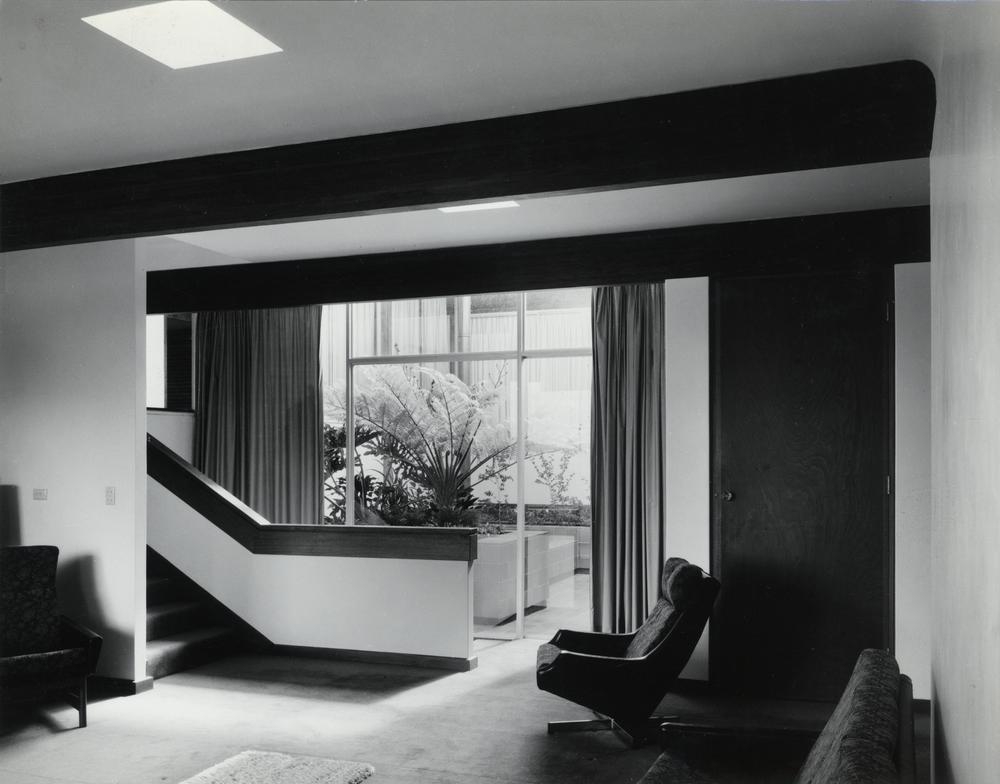 This is an image of ‘Interior of the Malouf residence in Holland Park, Queensland, 1969', showing an internal view of the heritage place from the sitting room, with staircase, to an external courtyard beyond.
