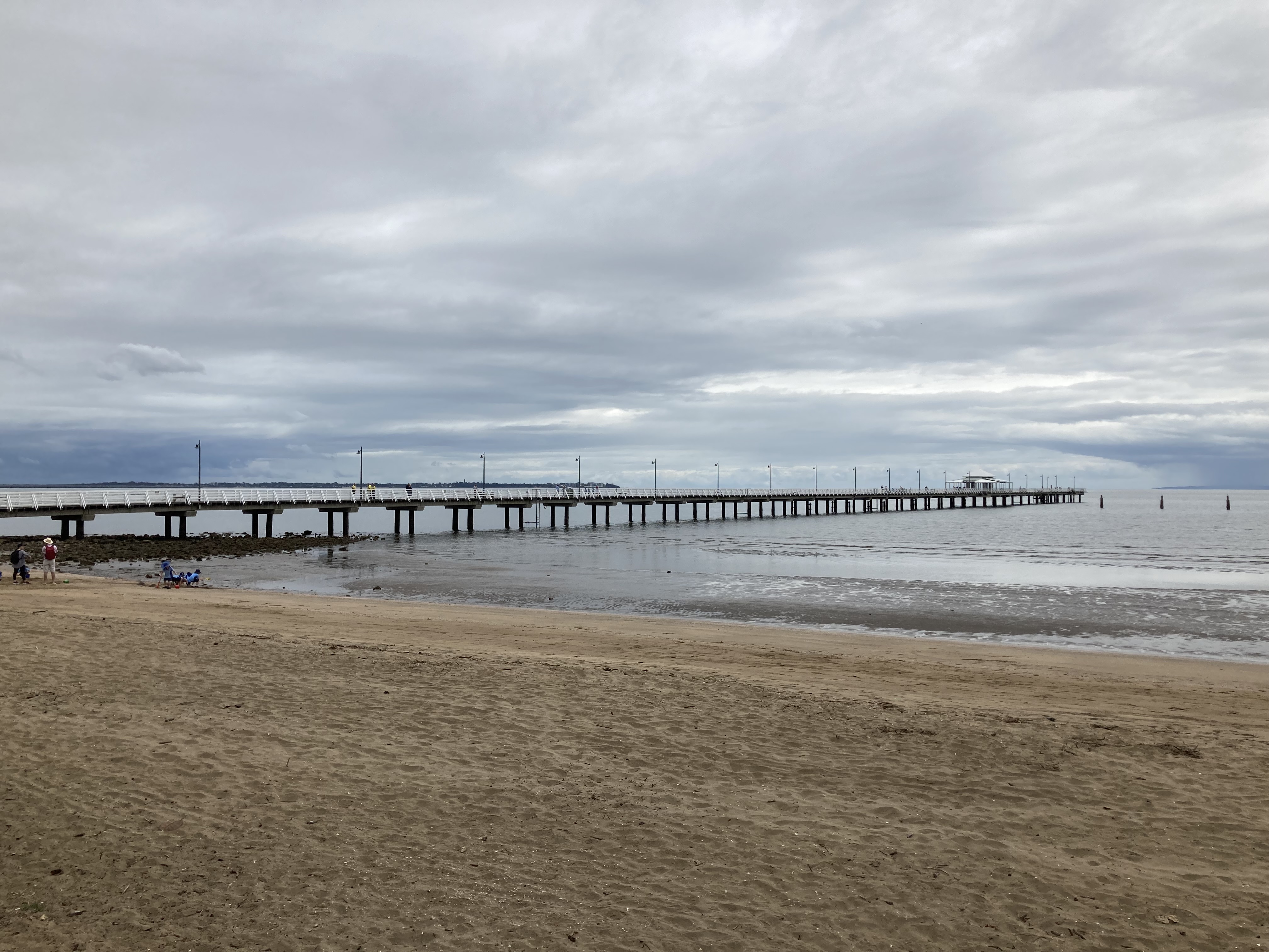 A long timber jetty, with a white roofed structure at the end, and a open sandy beach in the foreground