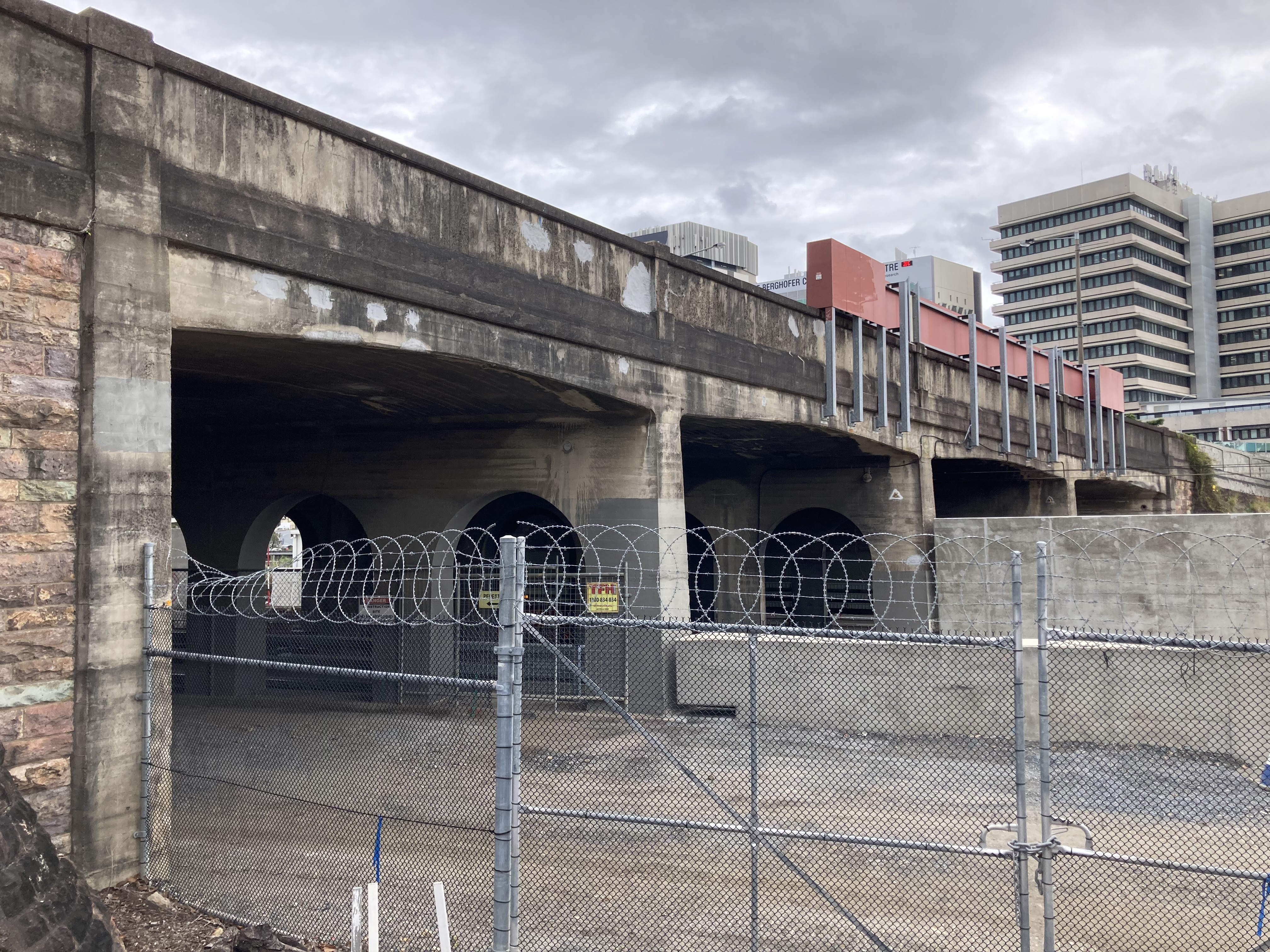 A concrete and brick bridge, with railway lines passing underneath. The four openings below the bridge are largely square, with slightly arched tops. A wire security fence, topped with spiral barbed wire, is in the foreground. Large hospital buildings are in the background.