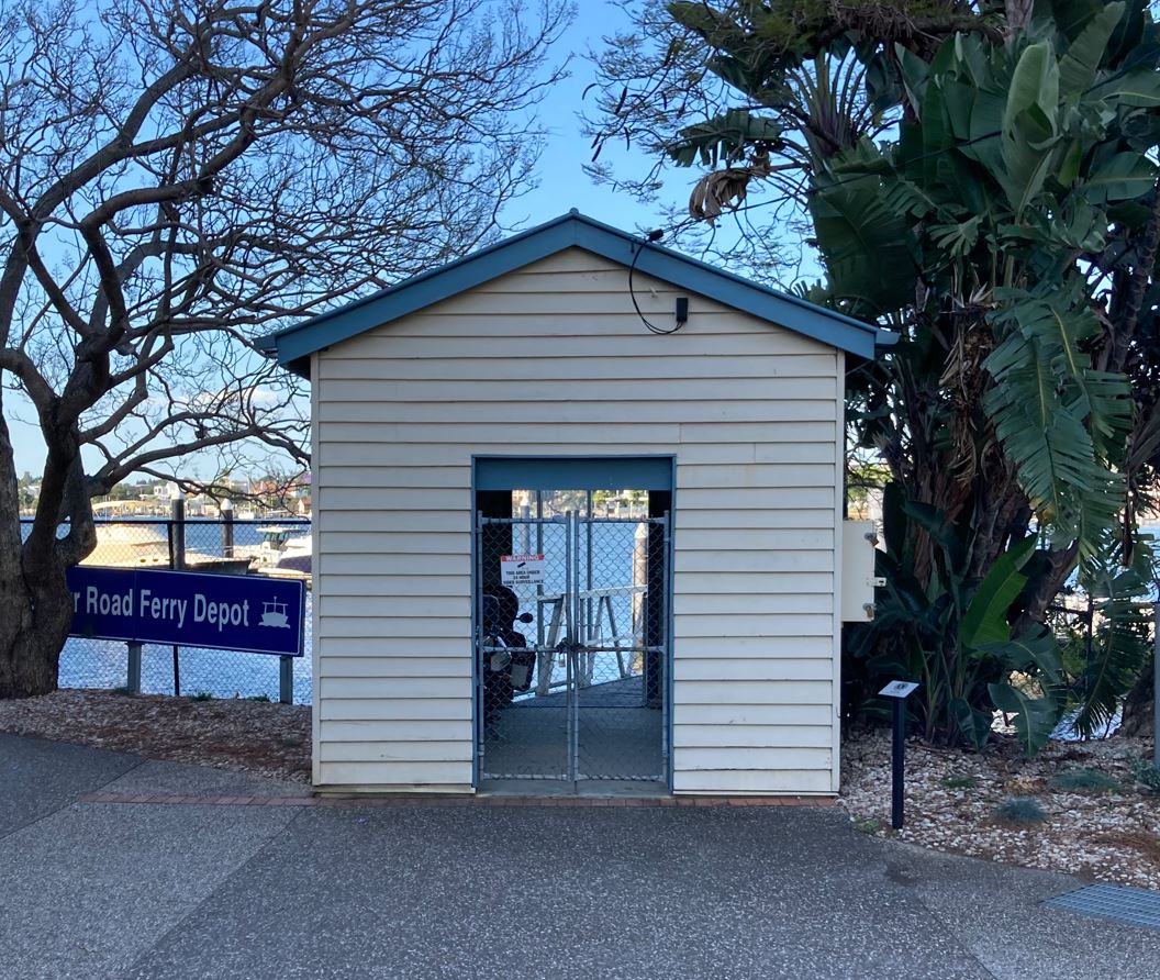 A single storey, timber ferry terminal, painted cream with blue trims. Mature trees are on either side of the structure, with a "Road Ferry Depot" sign to the left. The Brisbane river can be seen in the background.