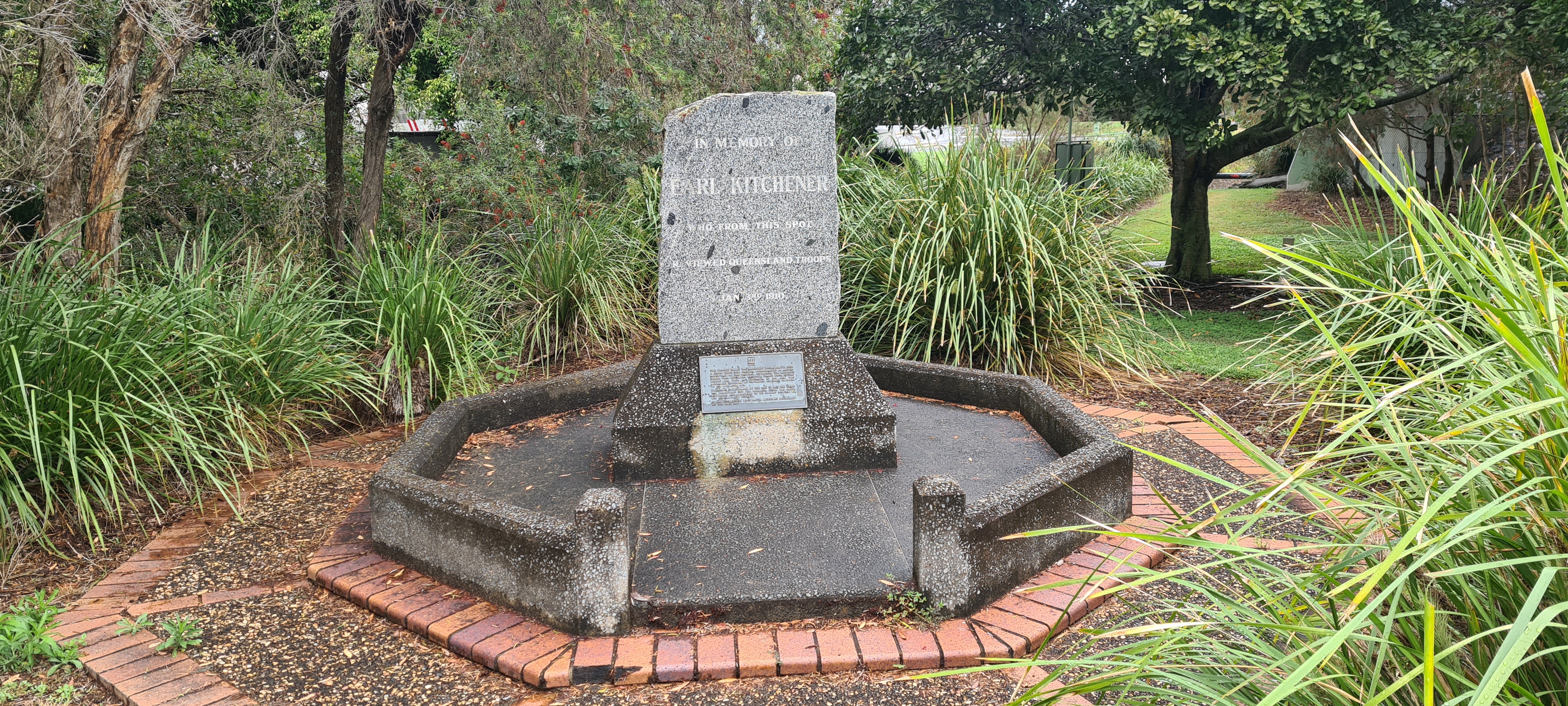 A stone and concrete memorial, bordered by brick and concrete, with vegetation in the background. 