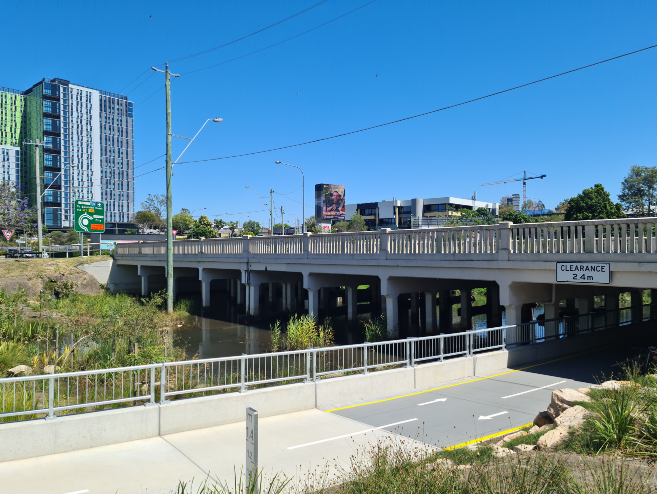 Burnett Swamp Bridge, a concrete bridge over a swampy area, with concrete footings faintly showing before the water. A concrete path with metal railings crosses under the bridge in the foreground. 