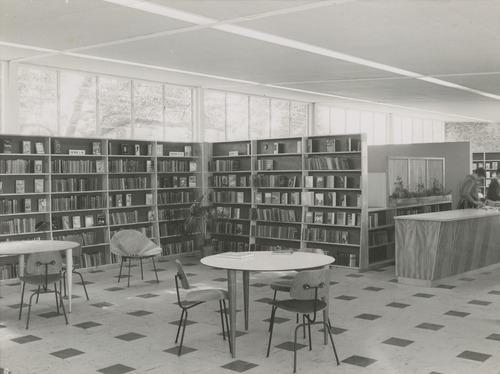 This is an image of ‘Interior view of the Annerley Library, Queensland, ca. 1957', showing a clerestory window-lit space, lined with bookshelves, a vinyl tiled floor with assorted tables and chairs and a library counter.