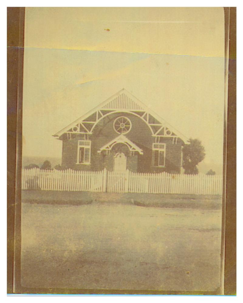 Single-story brick church building, with a white picket fence and gate at the front. A dirt road is in the foreground of the photograph